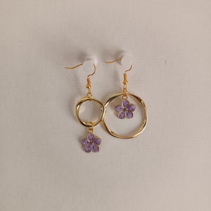 Puede incluir: Un par de pendientes dorados. Cada pendiente presenta un diseño de aro dorado con un pequeño dije de flor morada colgante. Los pendientes están suspendidos de ganchos dorados.