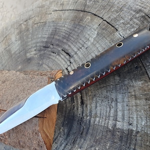May include: A handcrafted knife with a polished steel blade and a dark, textured handle. The handle features brass rivets and decorative stitching along the edges. The knife is designed for utility and display.