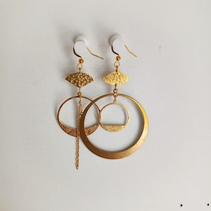 Op de afbeelding: Een paar goudkleurige oorbellen met een gelaagd ontwerp. Elke oorbel heeft een waaiervormige bovenkant, een kleine cirkel, een grotere cirkel en een bungelende ketting. De oorbellen hangen tegen een witte achtergrond.