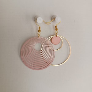 Puede incluir: Un par de pendientes asimétricos. Uno presenta un diseño circular grande, rosa claro y en capas. El otro tiene un aro dorado con un pequeño círculo rosa y detalles dorados. Los pendientes cuelgan de ganchos dorados.