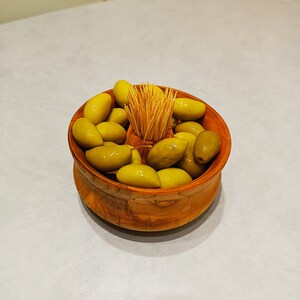Puede incluir: Un cuenco de madera lleno de aceitunas verdes y un manojo de palillos de dientes. El cuenco tiene un tono cálido de madera natural y está sobre una superficie clara. Las aceitunas son de color verde brillante.