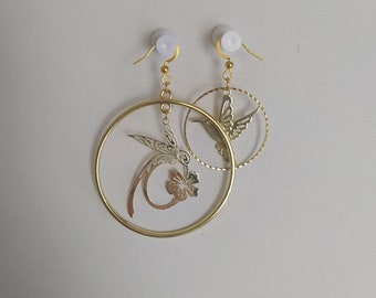 May include: A pair of gold-toned hoop earrings with intricate hummingbird and flower cutouts. The earrings feature a circular design with a smaller, textured hoop and are suspended from gold-colored ear wires. The background is a plain white surface.