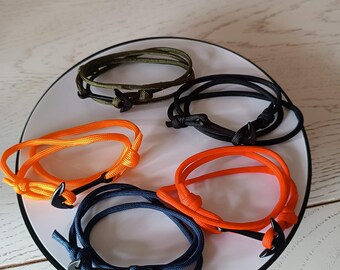 May include: Five rope bracelets in various colors: orange, navy blue, black, and olive green. Each bracelet features a black anchor-shaped clasp. The bracelets are displayed on a white plate.