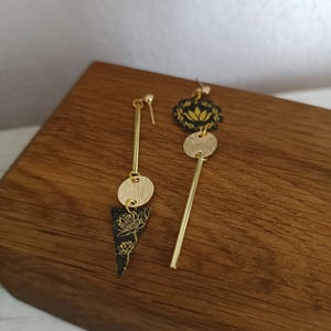 Pode incluir: Um par de brincos pendurados dourados. Um brinco apresenta uma barra dourada, um disco dourado e um triângulo preto com um desenho floral dourado. O outro brinco tem uma barra dourada, um disco dourado e um desenho floral preto.