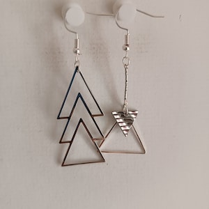 Op de afbeelding: Zilverkleurige geometrische oorbellen. Eén oorbel heeft drie gestapelde driehoeken, de andere een getextureerde driehoek en een verticale staaf. De oorbellen hangen aan zilveren haken.