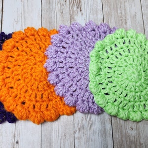 K&ouml;nnte beinhalten: Vier geh&auml;kelte Untersetzer in Lila, Orange, Lavendel und Gr&uuml;n. Die Untersetzer sind rund und haben ein strukturiertes Design.