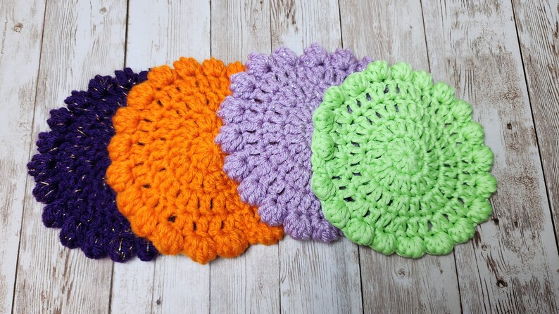 K&ouml;nnte beinhalten: Vier geh&auml;kelte Untersetzer in Lila, Orange, Lavendel und Gr&uuml;n. Die Untersetzer sind rund und haben ein strukturiertes Design.