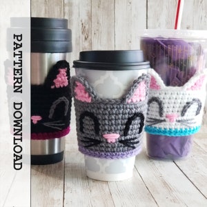 Op de afbeelding: Drie gehaakte koffiebekers met kattenmotief in grijs, zwart en wit. De bekers hebben roze oren en een zwarte neus. De bekers staan op verschillende koffiemokken.