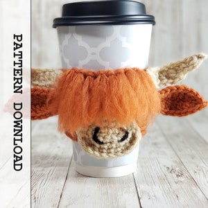 May include: A crocheted coffee cup cozy in the shape of a Highland cow. The cozy is brown and white with a fluffy orange mane. The cozy is on a white coffee cup.