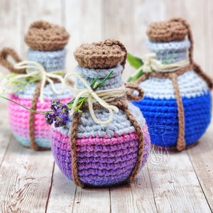 May include: Three crocheted potion bottles with cork tops. The bottles are decorated with different colored yarn, including pink, blue, purple, and gray. The bottles are tied with twine and have a rustic, handmade look.