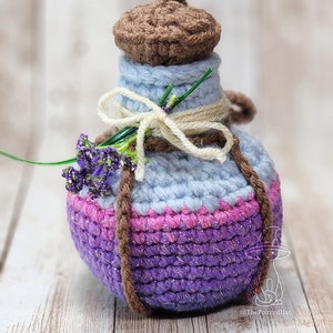 May include: A crocheted bottle with a brown lid, light blue body, pink and purple bottom, and a lavender flower on the side. The bottle is sitting on a wooden surface. @ThePointedHat