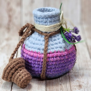May include: A crocheted potion bottle with a light blue, pink, and purple color gradient. The bottle has a brown cord and a brown crocheted stopper. The bottle is decorated with a small bunch of lavender.
