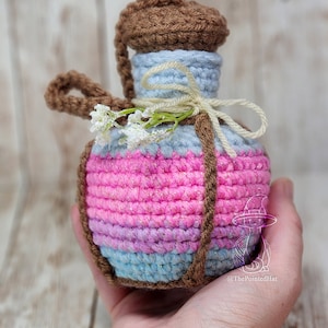 May include: A crocheted bottle with a brown cork top, a light blue body, and a pink, purple, and blue striped bottom. The bottle is decorated with a brown rope and a small sprig of white flowers.