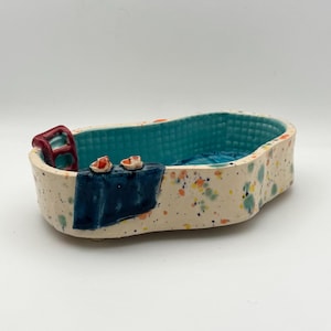 Puede incluir: Un plato de cerámica con forma de bañera con exterior moteado. El interior es turquesa con un patrón texturizado. Se incluye una pequeña escalera roja y una toalla azul con dos pequeños objetos naranjas.