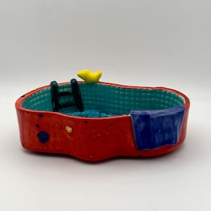 Könnte beinhalten: Eine handgefertigte Miniatur-Keramik-Pool. Das Äußere ist rot, das Innere türkis und es gibt ein blaues Handtuch. Eine kleine schwarze Leiter und eine gelbe Quietscheente sind ebenfalls vorhanden. Der Pool ist ein dekorativer Artikel.