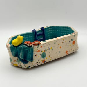 Könnte beinhalten: Eine skurrile Keramikbadewanne mit türkisfarbenem Innenraum und einer Leiter. Die Außenseite ist mit bunten Punkten gesprenkelt. Eine gelbe Ente, ein türkisfarbenes Handtuch und winzige rote Sandalen sind an der Seite platziert. Ein einzigartiges Dekorationsstück.