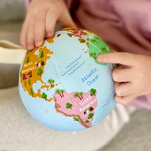 May include: A soft fabric globe toy with a light blue background and colourful illustrations of continents and countries. The globe has a beige handle and is being held by a child. The words "North America", "Atlantic Ocean", and "South America" are visible.