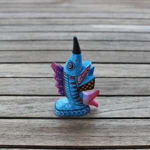 Könnte beinhalten: Eine handbemalte Holzfigur eines Fisches mit blauen, rosa, gelben und schwarzen Details. Der Fisch hat eine lange, spitze Schnauze und ein großes, rundes Auge.