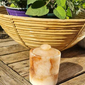 May include: A cylindrical, lidded container made of pink and brown-streaked stone. The container sits on a wooden surface, with a woven basket and potted plants in the background. The lid has a small, rounded knob.