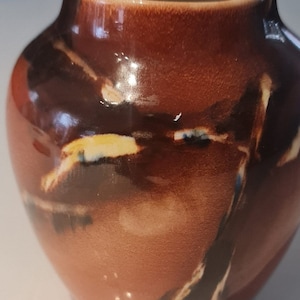 May include: A small, brown ceramic vase with a glossy finish. The vase has a unique, abstract design with black, white, and yellow accents.