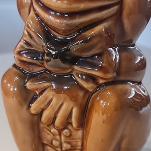 Peut inclure: Une figurine de grenouille en céramique brune avec un nœud papillon. La grenouille est assise sur une base avec le mot "Fred" écrit dessus.