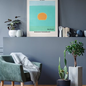 May include: A framed print of a sunset painting by Andy Warhol. The painting is a simple design of a yellow sun on a blue and green background. The text "Andy Warhol" and "Museum of Modern Art" are printed below the painting.