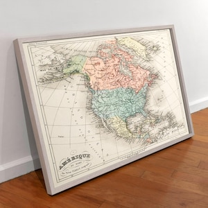 May include: A framed vintage map of North America, with detailed geographical information and historical cartography. The map is in a light-coloured wooden frame, with text in French. The map's colours include shades of green, pink, and yellow.