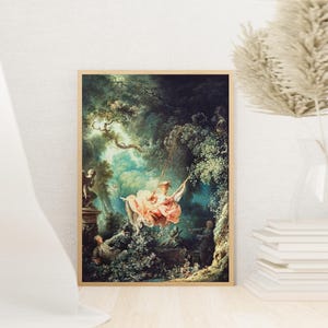 May include: A framed print of a Rococo painting showing a woman on a swing in a verdant forest. The woman is dressed in a pink gown and hat. Other figures are present, and the artwork is in a light brown wooden frame.