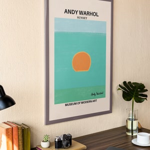May include: A framed print of a sunset painting by Andy Warhol. The painting is a simple design of a large orange sun setting over a turquoise ocean. The text "Andy Warhol" and "Museum of Modern Art" are printed below the painting.