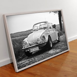 May include: Black and white photograph of a classic Volkswagen Beetle convertible with a number plate that reads "VER 1976 H". The car is parked on a cobbled road with trees and grass in the background.