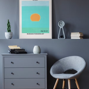 May include: A gray dresser with three drawers and black knobs. A vintage typewriter sits on top of the dresser. A gray armchair with wooden legs sits in front of the dresser. A framed poster with the text "ANDY WARHOL SUNSET" and "MUSEUM OF MODERN ART" hangs on a gray wall. A small potted plant and a clock are on a shelf above the dresser.