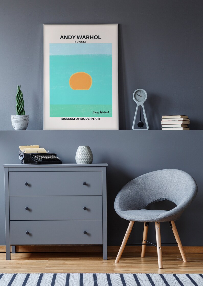 May include: A gray dresser with three drawers and black knobs. A vintage typewriter sits on top of the dresser. A gray armchair with wooden legs sits in front of the dresser. A framed poster with the text "ANDY WARHOL SUNSET" and "MUSEUM OF MODERN ART" hangs on a gray wall. A small potted plant and a clock are on a shelf above the dresser.