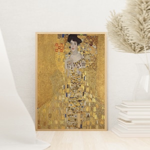 May include: Framed print of a Gustav Klimt painting. The artwork features a woman in an ornate gold dress with geometric patterns. The background is also gold, with intricate details. The frame is a light wood color.