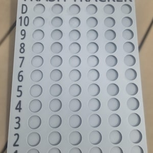 May include: A rectangular light gray plastic tracker board with the text TRASH TRACKER at the top. The board features a grid of ten rows and seven columns of circular indentations, with the numbers 1 through 10 and the letter D printed vertically.