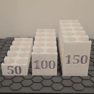 May include: Three white, geometric, stepped structures with the numbers 50, 100, and 150 printed on them. The structures have a honeycomb-like pattern on top. They are placed on a gray, hexagonal patterned surface.
