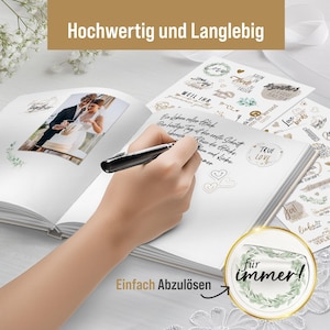 May include: A white wedding guest book with a photo of a bride and groom on the cover. The book has a gold inscription that reads "Hochwertig und Langlebig" and a sticker that says "True Love". The book is open to a blank page with a hand writing on it. The book is on a white surface with a green and white sticker that says "F&uuml;r immer!" and the text "Einfach Abzul&ouml;sen".