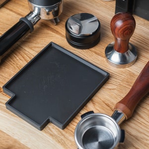 Puede incluir: Un conjunto de herramientas para hacer café, que incluye una esterilla de manipulación negra, un pisón con mango de madera, un portafiltro y un pisón negro. Los artículos están dispuestos sobre una superficie de madera clara, lo que sugiere una configuración de preparación de café.