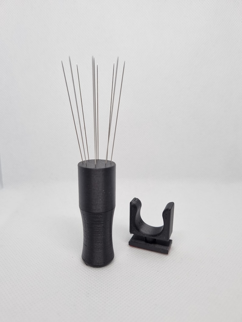 K&ouml;nnte beinhalten: Ein schwarzes Kaffee-Verteilerwerkzeug mit d&uuml;nnen Metallnadeln und einem passenden Halter. Das Werkzeug hat einen zylindrischen Griff mit strukturierter Oberfl&auml;che. Der Halter ist ein kleines, gebogenes Teil.