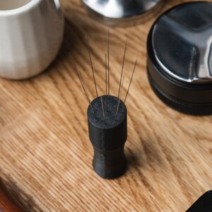 K&ouml;nnte beinhalten: Ein schwarzes Kaffee-Verteilungswerkzeug mit mehreren d&uuml;nnen Nadeln, die aus der Oberseite herausragen. Das Werkzeug ist zylindrisch mit einem ausgestellten Boden. Weitere Kaffeezubereitungsutensilien, darunter ein Tamper und eine kleine wei&szlig;e Keramiktasse, sind sichtbar.
