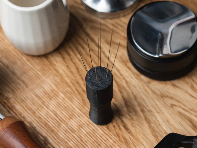 K&ouml;nnte beinhalten: Ein schwarzes Kaffee-Verteilungswerkzeug mit mehreren d&uuml;nnen Nadeln, die aus der Oberseite herausragen. Das Werkzeug ist zylindrisch mit einem ausgestellten Boden. Weitere Kaffeezubereitungsutensilien, darunter ein Tamper und eine kleine wei&szlig;e Keramiktasse, sind sichtbar.