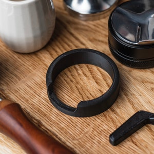 Puede incluir: Un anillo negro para accesorios de café sobre una superficie de madera, junto con un portafiltro, un tamper y otras herramientas para hacer café. El anillo tiene un diseño curvo con un recorte.