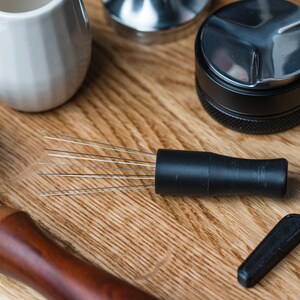 K&ouml;nnte beinhalten: Kaffeezubereitungsutensilien auf einer Holzoberfl&auml;che. Abgebildet sind ein schwarzer Kaffee-Verteiler mit Nadeln, ein Tamper, ein wei&szlig;er Keramik-Krug und ein Siebtr&auml;ger mit Holzgriff.