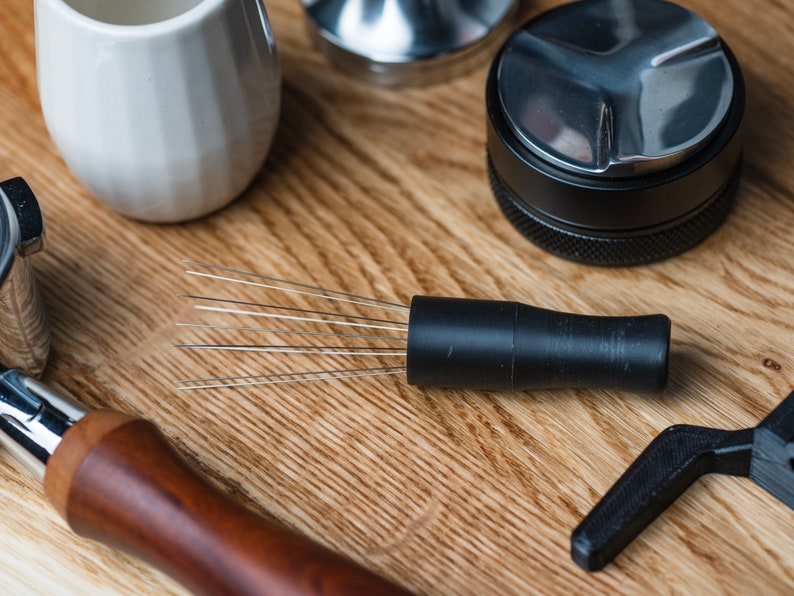 K&ouml;nnte beinhalten: Kaffeezubereitungsutensilien auf einer Holzoberfl&auml;che. Abgebildet sind ein schwarzer Kaffee-Verteiler mit Nadeln, ein Tamper, ein wei&szlig;er Keramik-Krug und ein Siebtr&auml;ger mit Holzgriff.