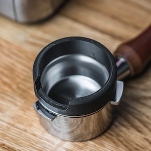 May include: A stainless steel portafilter with a black rim and a wooden handle. The portafilter is designed for espresso machines and sits on a wooden surface. The image is a close-up shot, highlighting the details of the coffee-making tool.