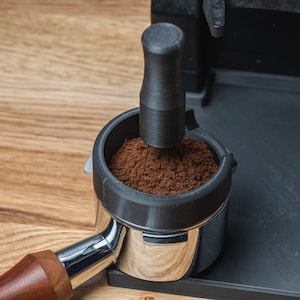 K&ouml;nnte beinhalten: Nahaufnahme eines Kaffeefilterhalters mit frisch gemahlenem Kaffee, der angedr&uuml;ckt wird. Der Filterhalter ist aus Edelstahl mit einem Holzgriff. Das Tamperwerkzeug ist schwarz. Der Hintergrund ist eine hellbraune Holzoberfl&auml;che.