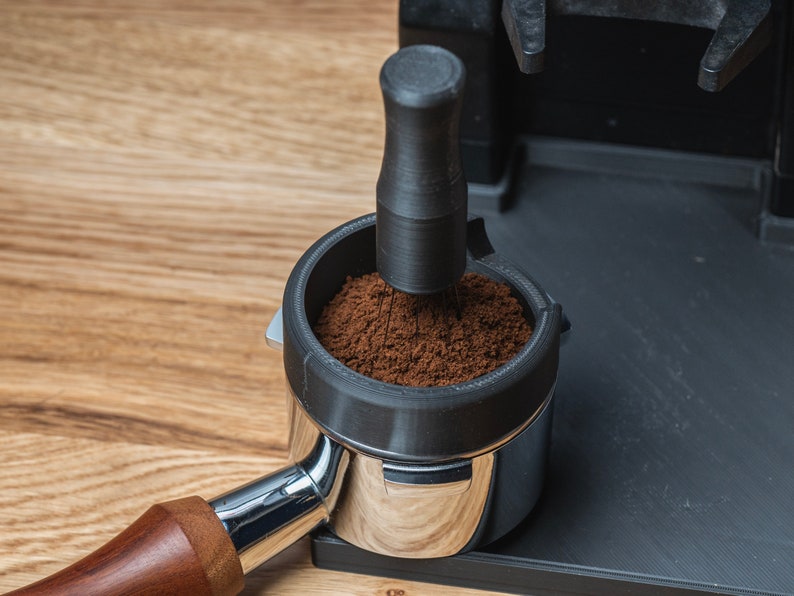 K&ouml;nnte beinhalten: Nahaufnahme eines Kaffeefilterhalters mit frisch gemahlenem Kaffee, der angedr&uuml;ckt wird. Der Filterhalter ist aus Edelstahl mit einem Holzgriff. Das Tamperwerkzeug ist schwarz. Der Hintergrund ist eine hellbraune Holzoberfl&auml;che.