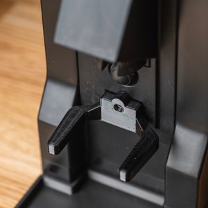 Op de afbeelding: Close-up van een zwarte koffiemachine met een 3D-geprint onderdeel. Het onderdeel heeft twee armen die naar beneden uitsteken, ontworpen om een kopje vast te houden. Het ontwerp van de machine is modern en strak, met focus op functionaliteit.