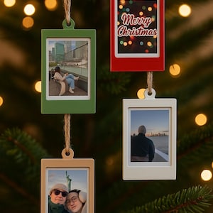 Könnte beinhalten: Vier Weihnachtsschmuckstücke in Form von Polaroidfotos. Eines der roten Schmuckstücke trägt die Aufschrift "Merry Christmas". Die anderen drei zeigen Fotos von Menschen und Orten. Jedes Schmuckstück hat eine andere Farbe: grün, beige und weiß.