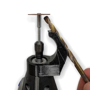 Könnte beinhalten: Nahaufnahme eines schwarzen Rotationswerkzeugs mit einer kleinen, runden, braunen Schleifscheibe, die an der Spindel befestigt ist. Ein goldener Bohrer wird in der Hand gehalten und ist bereit, in die Spannzange des Werkzeugs eingesetzt zu werden.