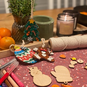 May include: A festive scene with Christmas ornaments, including a tree, gingerbread man, and bauble. Wooden snowman and mitten cutouts are also present, along with markers and twine. The ornaments are on a red patterned surface, suggesting a craft or holiday theme.